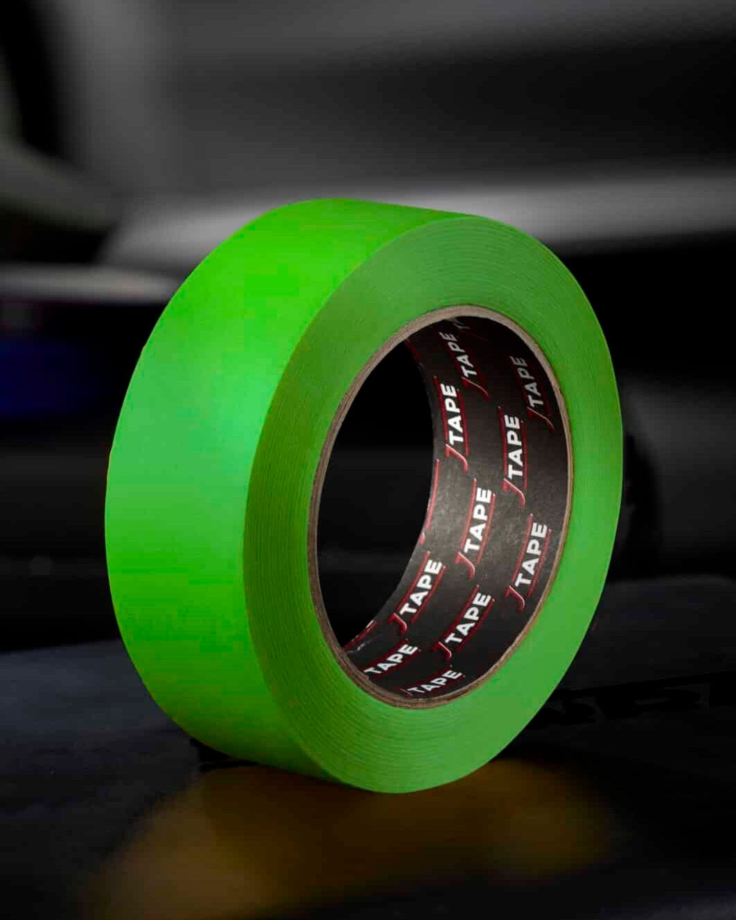 A roll of bright green tape, standing upright on a dark surface, with a blurred background that includes another roll of tape and a car door.